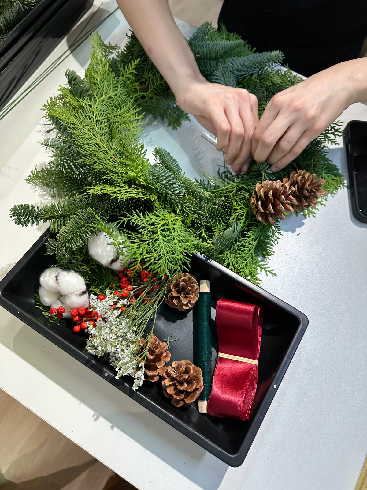 Hands arranging fresh pine onto a wreath, with pinecones and red ribbon in a tray