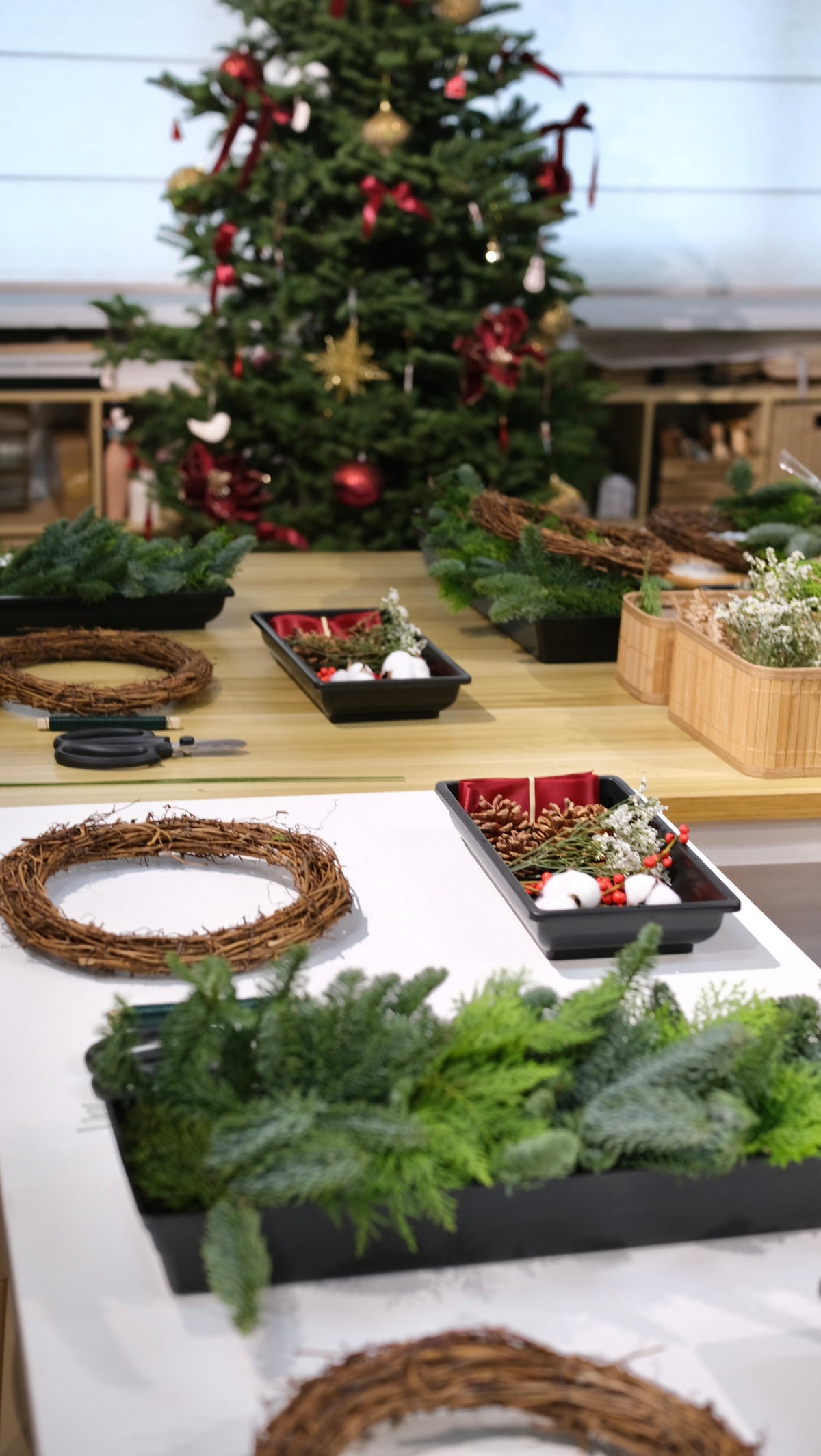 Workshop table set with rattan wreath bases, trays of fresh pine and red decorations in front of a Christmas tree