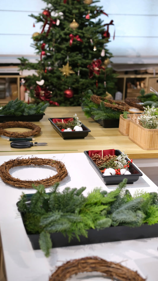 Workshop table set with rattan wreath bases, trays of fresh pine and red decorations in front of a Christmas tree