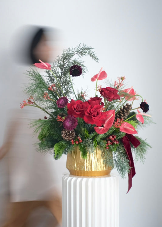 Crimson Noel Christmas centrepiece with red roses, pink anthuriums, pine foliage, berries and pinecones in a gold vase