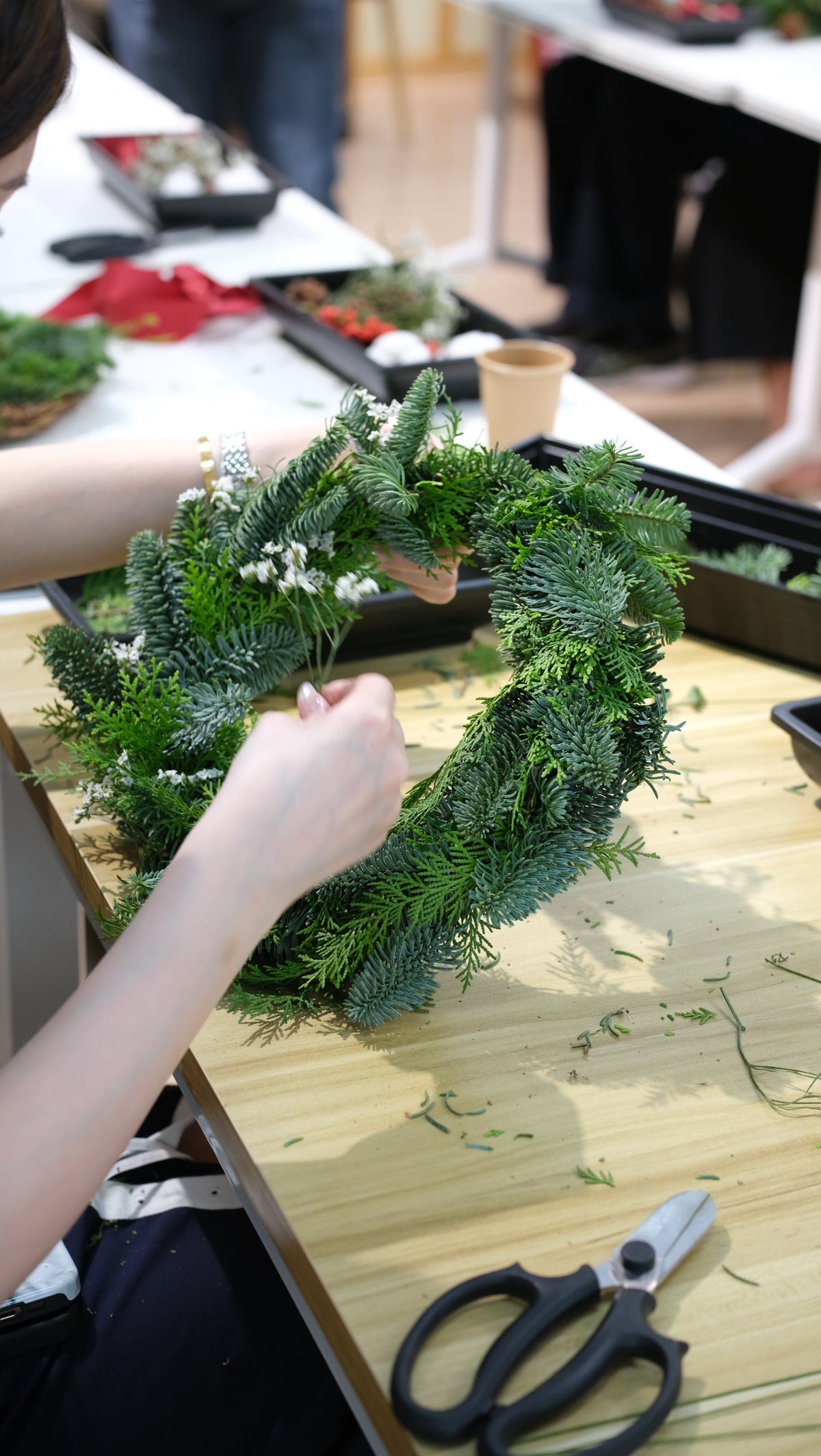 Participant tying fresh pine foliage onto a wreath base with floral scissors on the table