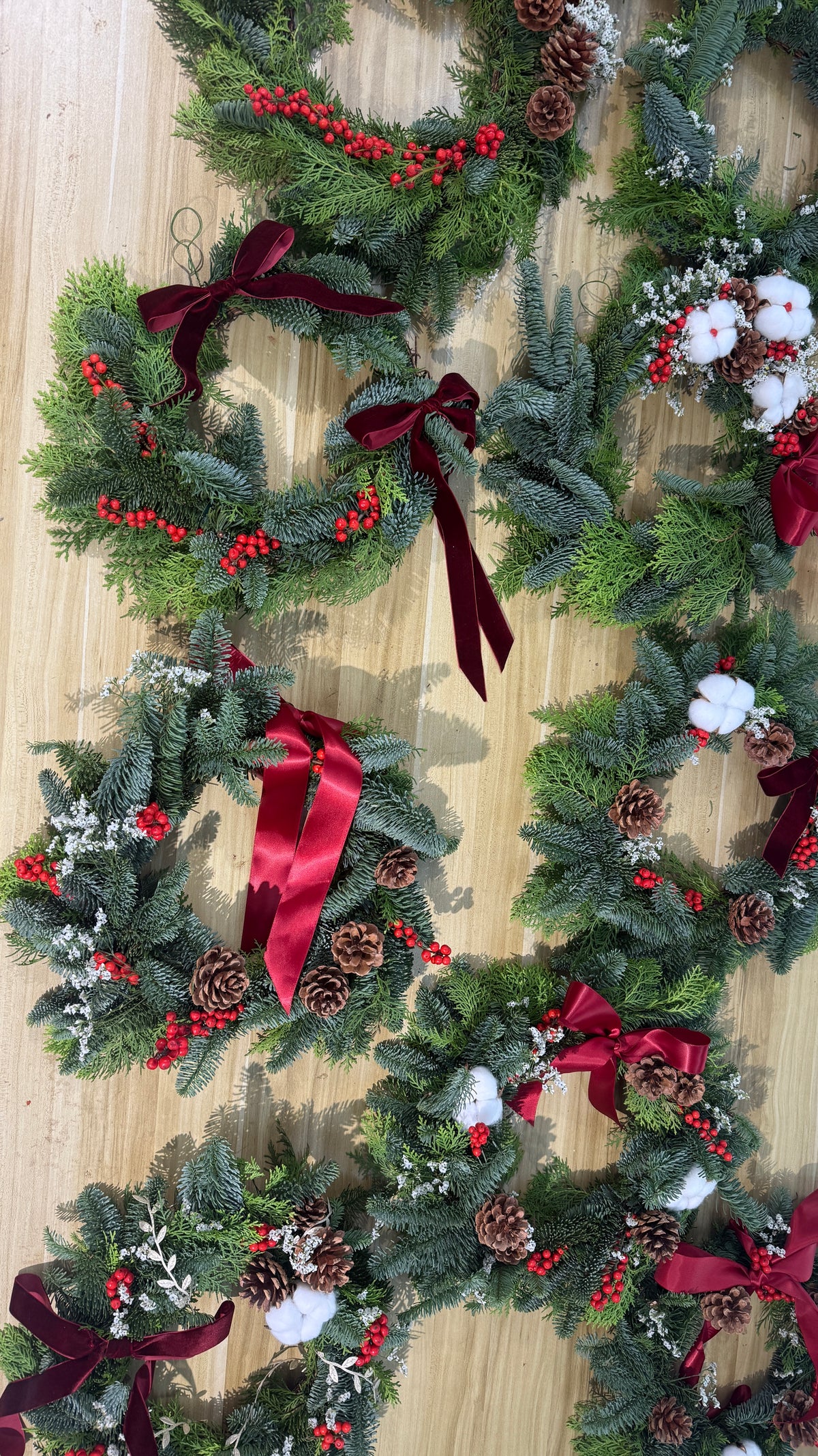 Group of finished fresh Christmas wreaths with red ribbons and cotton flowers laid on a wooden table