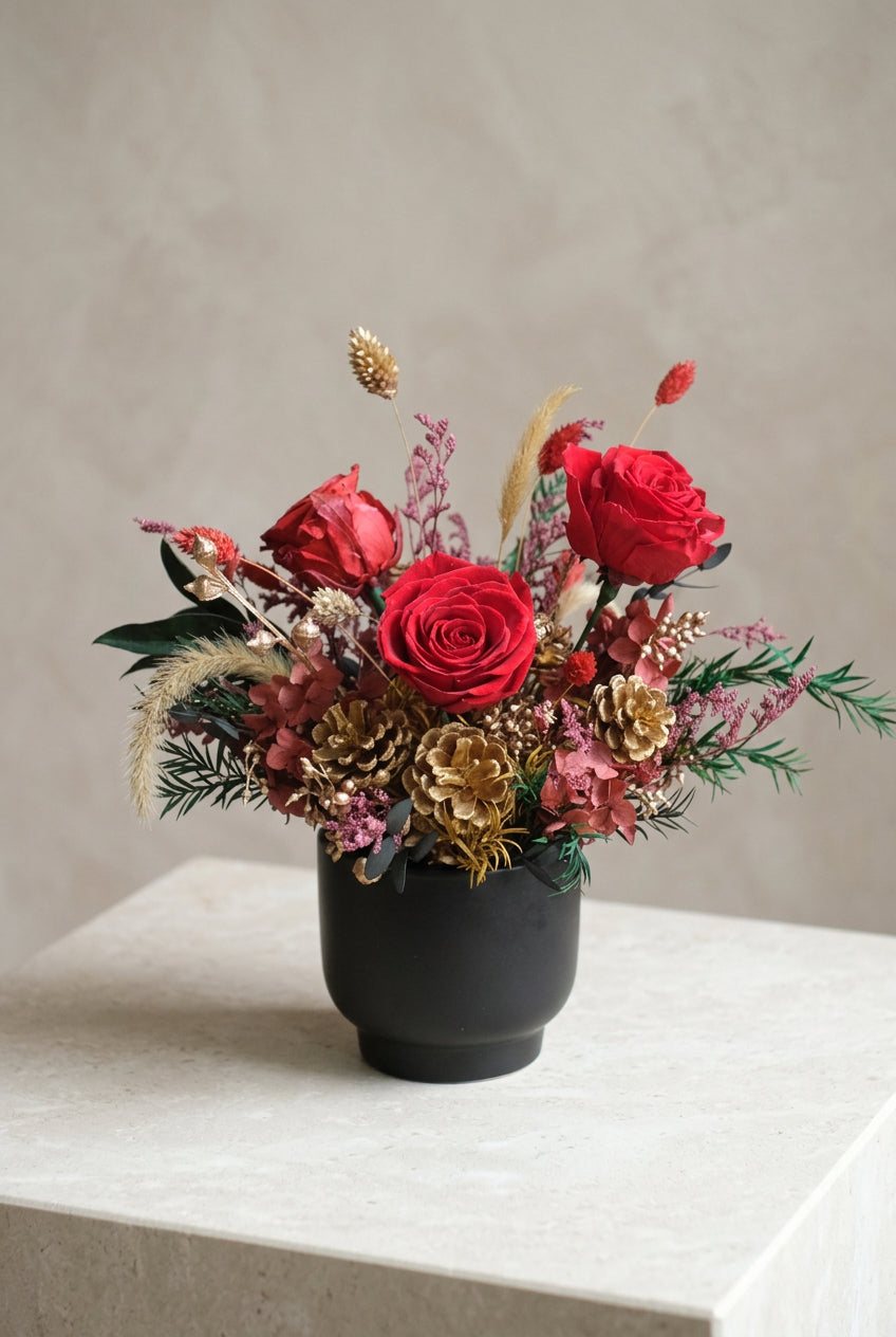 Holiday Enchantment preserved Christmas arrangement with red roses, gold pinecones and burgundy hydrangea in a matte black vase