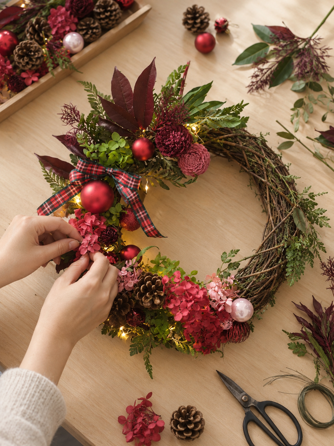 Hands arranging red preserved flowers and berries onto a rattan wreath base at a Christmas workshop